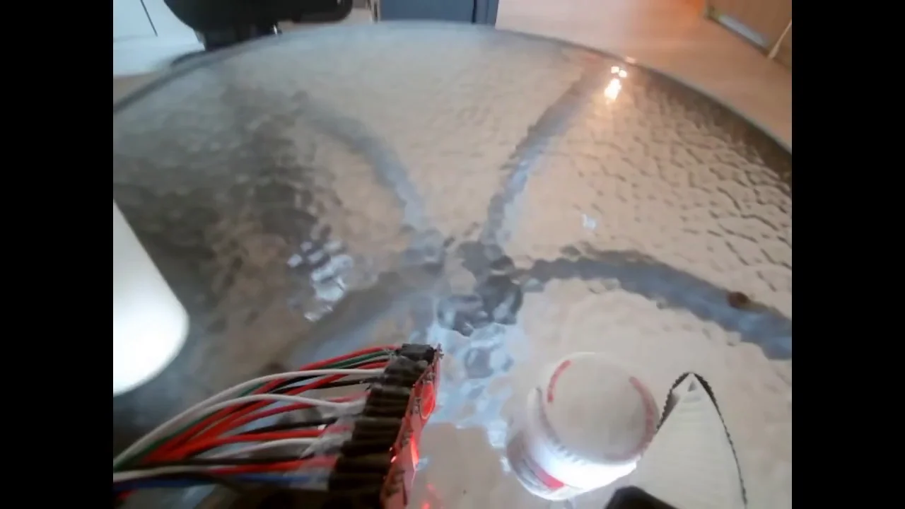 Wrist-camera recording: a robot gripper picks up a pill bottle from a glass table, carries it, and drops it into a paper cup.